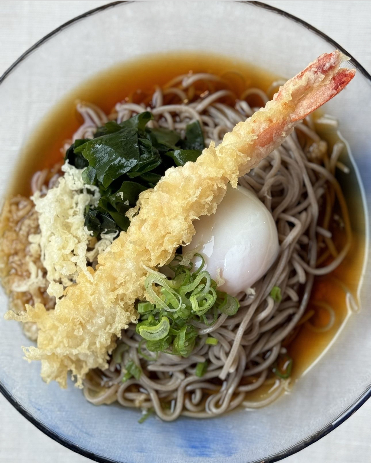 SOBA: pasta di grano saraceno, arricchita da una deliziosa tempura di gambero, uovo bollito, alghe wakame, cipollotti freschi e un tocco di sesamo croccante.